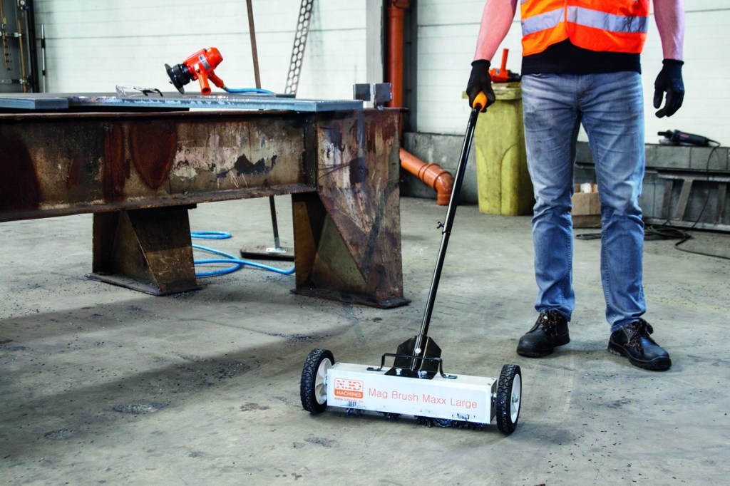A worker in a safety vest using a magnetic broom to clean a workshop floor, with a metal table and tools visible in the background.